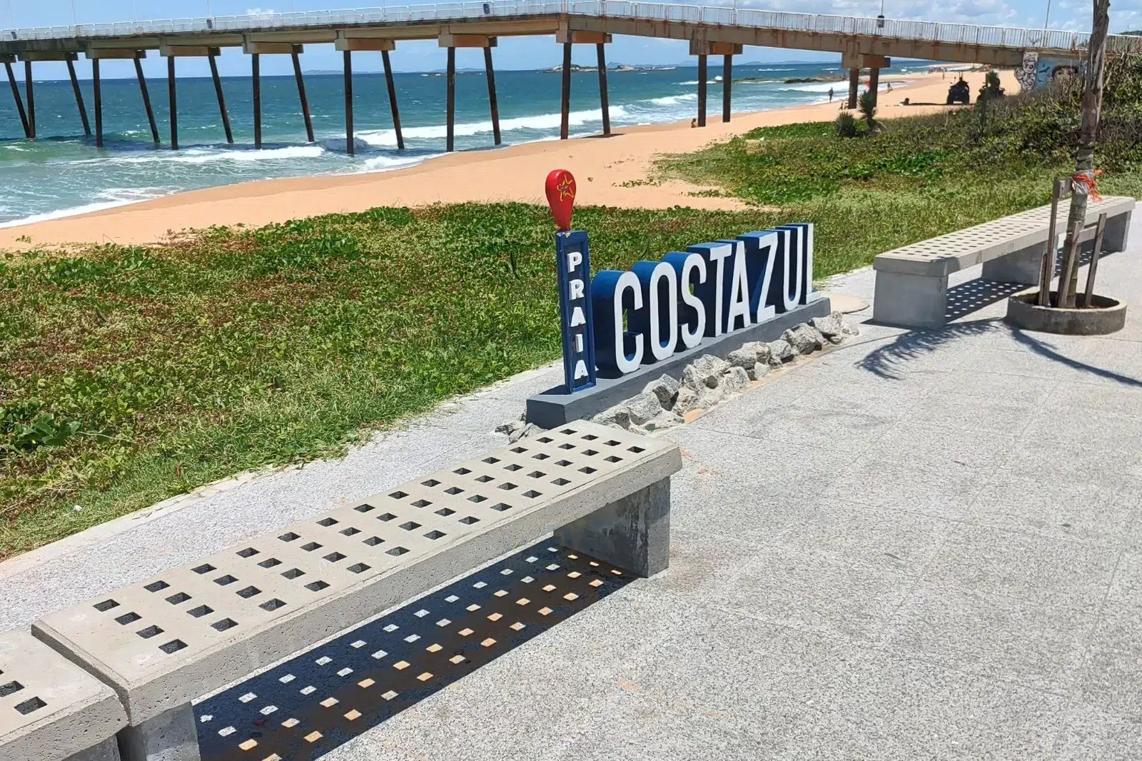 Bancos de concreto pré-moldado com assento perfurado e design vazado em uma orla à beira-mar, perto de um píer longo e da placa de identificação da Praia Costa Azul.